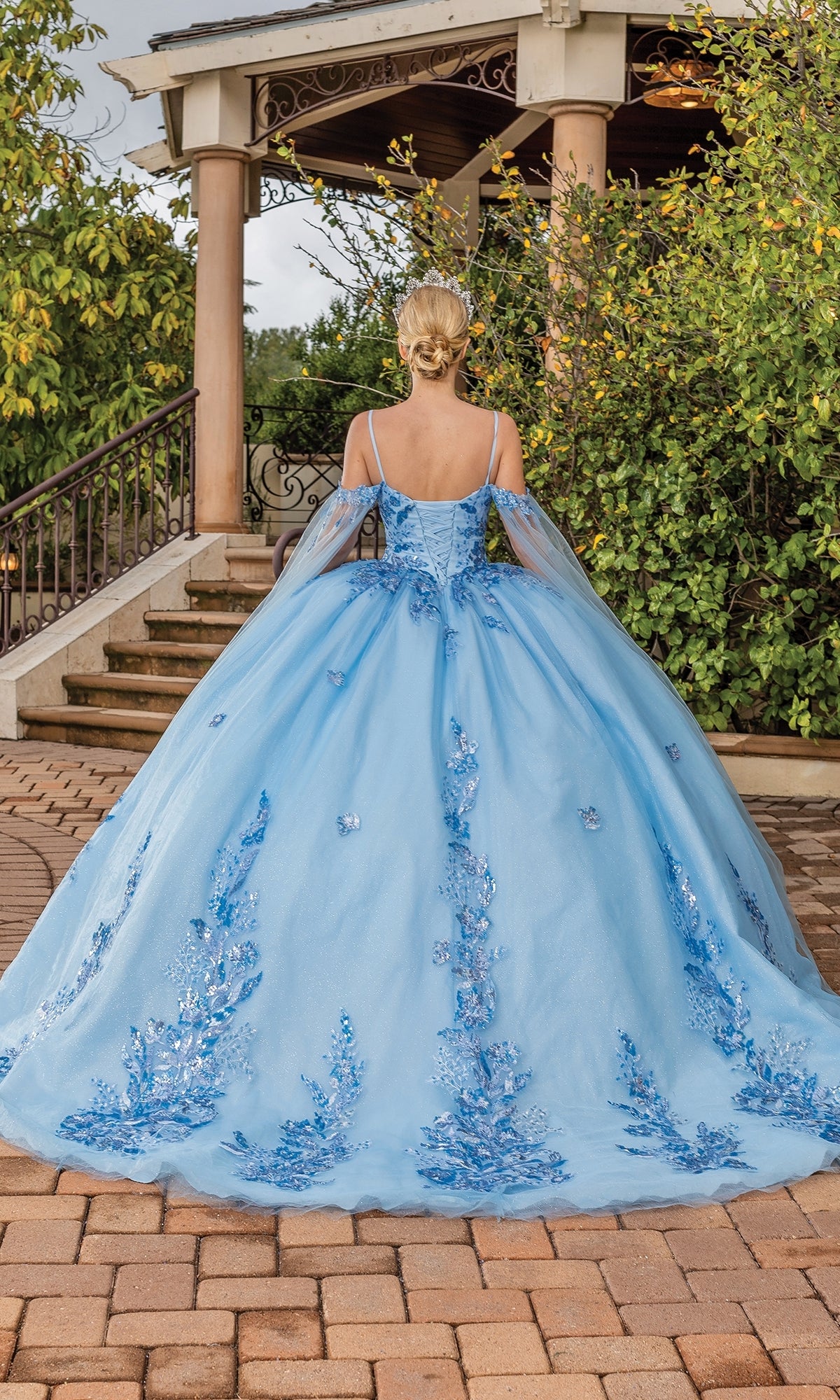 A person stands outdoors in front of a gazebo, facing away from the camera, wearing the Quinceanera Dress 1865 By Dancing Queen—an a-line ball gown with floral embellishments and off-the-shoulder sleeves.